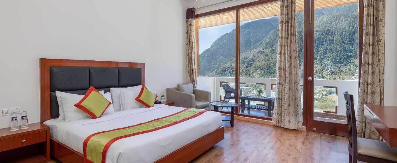 The image shows a hotel room with a large bed, a sitting area with a sofa and coffee table, and a large window with a view  | DLS Serenity Resort and Spa